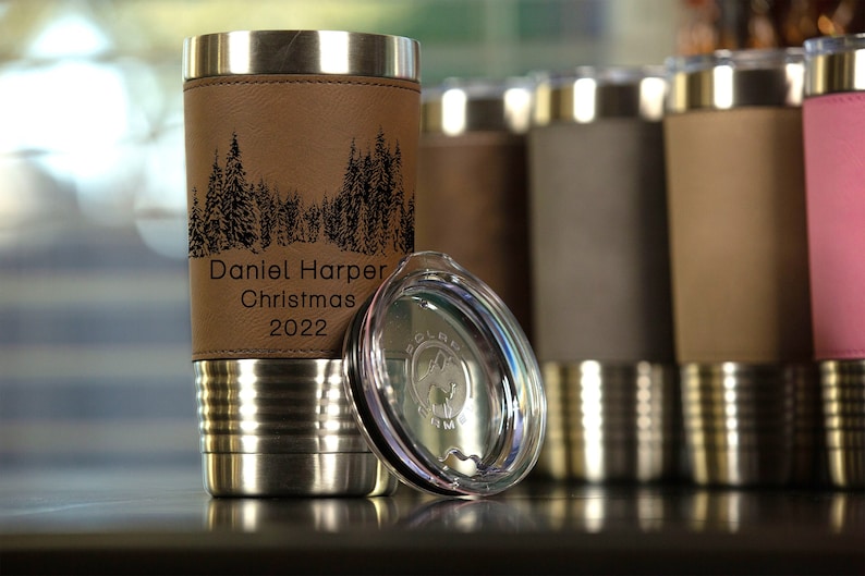 May include: A stainless steel tumbler with a brown leatherette wrap. The tumbler has a black laser engraved design of a forest with trees and the text "Daniel Harper Christmas 2022". The tumbler comes with a clear lid.