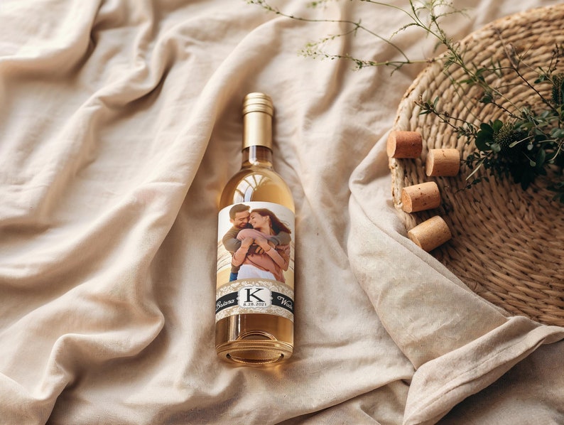 Pode incluir: Uma garrafa de vinho branco de vidro transparente com um r&oacute;tulo personalizado que mostra uma foto de um casal se abra&ccedil;ando. O r&oacute;tulo tem um design preto e dourado com a letra "K" no centro. A garrafa est&aacute; sobre uma superf&iacute;cie de tecido branco.