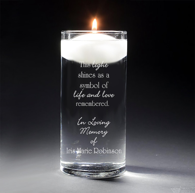 May include: A clear glass cylinder vase with a white floating candle and an engraved message: "This light shines as a symbol of life and love remembered. In Loving Memory of Iris Marie Robinson."