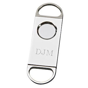 May include: A shiny, silver cigar cutter with a circular blade and a rectangular body. The initials "DJM" are engraved on the front. The cutter has a loop at the top and bottom for easy handling.