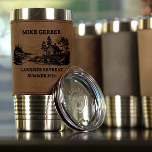 May include: A stainless steel tumbler with a brown leather wrap. The tumbler has a lid and is engraved with the text "Mike Gerber Lakeside Retreat Summer 2024".