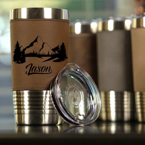 May include: A stainless steel travel mug with a brown leatherette sleeve. The sleeve has a mountain scene and the name "Jason" engraved on it. The mug comes with a clear lid.