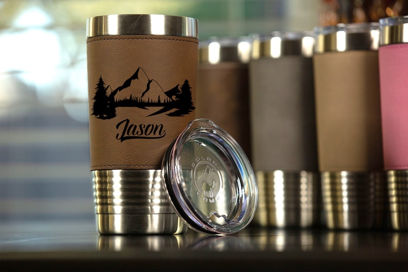 May include: A stainless steel travel mug with a brown leatherette sleeve. The sleeve has a mountain scene and the name "Jason" engraved on it. The mug comes with a clear lid.