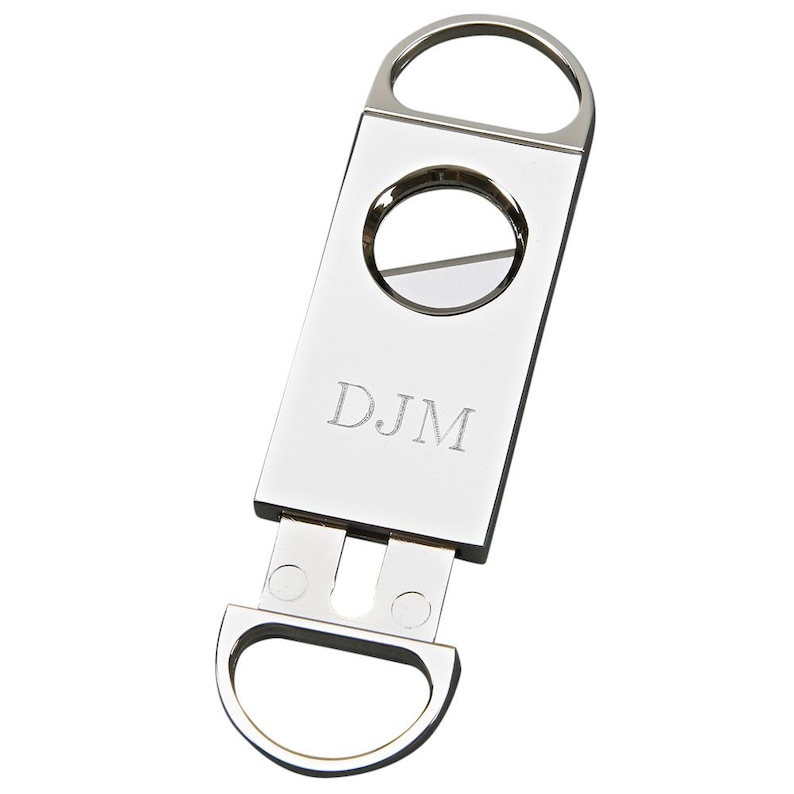 May include: A shiny, silver cigar cutter with a circular blade and a rectangular body. The initials "DJM" are engraved on the front. The cutter has a ring at the top and a curved handle at the bottom.