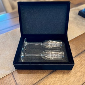 Op de afbeelding: Twee champagnefluiten in een zwarte fluwelen doos. De fluiten zijn gegraveerd met de woorden "Cheers to our forever" en "Happily ever after".