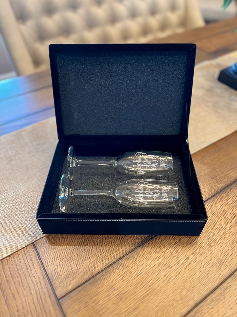 Op de afbeelding: Twee champagnefluiten in een zwarte fluwelen doos. De fluiten zijn gegraveerd met de woorden "Cheers to our forever" en "Happily ever after".