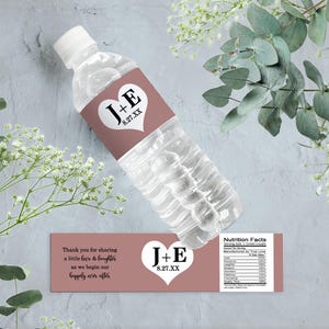 Op de afbeelding: Waterfleslabel met een roze achtergrond en een wit hart met de tekst "J+E 8.27.XX" in zwart. Het label bevat ook de tekst "Bedankt voor het delen van een beetje liefde en gelach terwijl we beginnen aan ons geluk voor altijd." en "Voedingswaarde".