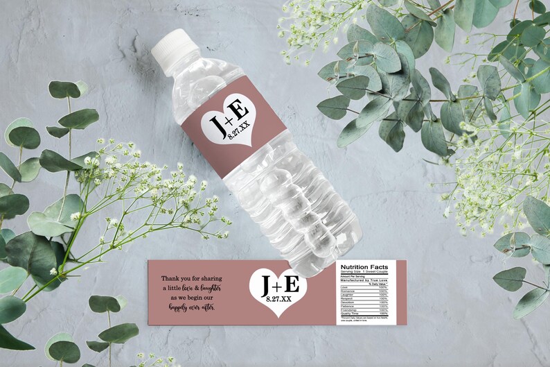 Op de afbeelding: Waterfleslabel met een roze achtergrond en een wit hart met de tekst "J+E 8.27.XX" in zwart. Het label bevat ook de tekst "Bedankt voor het delen van een beetje liefde en gelach terwijl we beginnen aan ons geluk voor altijd." en "Voedingswaarde".