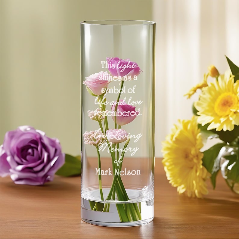 May include: A clear glass vase with a quote engraved on the side: "This light shines as a symbol of life and love remembered. In loving memory of Mark Nelson". The vase is filled with pink flowers and green stems.