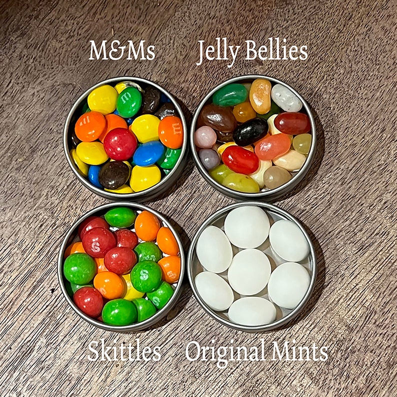 May include: Four small round metal tins filled with different types of candy. The tins are arranged in a square pattern on a wooden surface. The candies are M&Ms, Jelly Bellies, Skittles, and Original Mints.