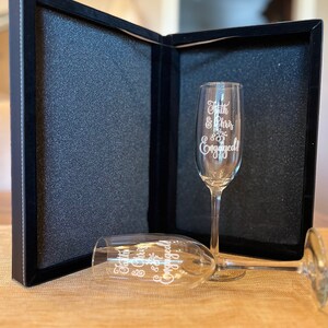 Op de afbeelding: Twee champagnefluiten met de tekst "Faith & Chris are Engaged!" erop gegraveerd. De fluiten zitten in een zwarte fluwelen doos.
