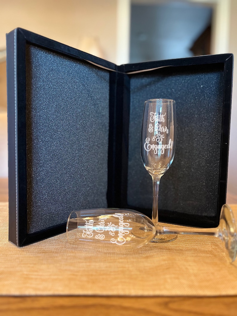 Op de afbeelding: Twee champagnefluiten met de tekst "Faith & Chris are Engaged!" erop gegraveerd. De fluiten zitten in een zwarte fluwelen doos.