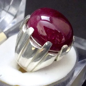 May include: A silver ring featuring a large, oval-shaped, deep red ruby gemstone.