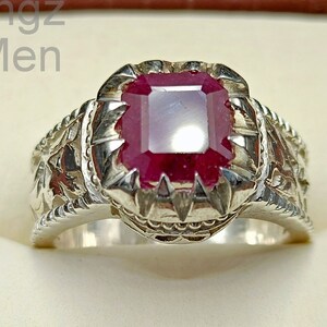 May include: A silver ring with a large, square-cut ruby gemstone. The ring has intricate floral and leaf detailing on the band and around the gemstone setting. The ruby is a deep red color.
