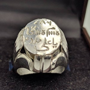 Op de afbeelding: Een zilveren ring met een groot ovaalvormig bovenstuk. De bovenkant van de ring is gegraveerd met Arabische tekst.