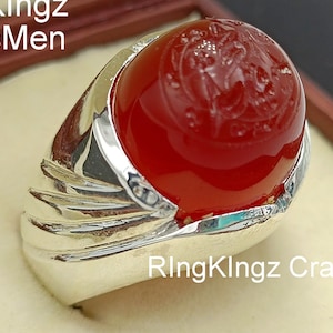 May include: A red carnelian gemstone ring set in a silver band. The ring features a detailed design on the gemstone and a textured band.