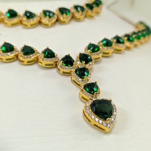 Op de afbeelding: Een gouden ketting met een hanger met een grote groene hartvormige edelsteen omringd door kleinere heldere edelstenen. De ketting is gemaakt van een ketting van kleinere groene hartvormige edelstenen.