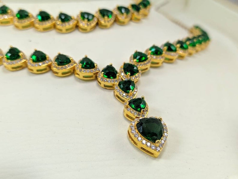 Op de afbeelding: Een gouden ketting met een hanger met een grote groene hartvormige edelsteen omringd door kleinere heldere edelstenen. De ketting is gemaakt van een ketting van kleinere groene hartvormige edelstenen.