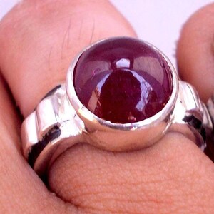 Pode incluir: Um anel de prata com uma grande pedra preciosa redonda e vermelha escura. A pedra preciosa está engastada numa banda de prata polida com um design simples. O anel é mostrado num dedo.