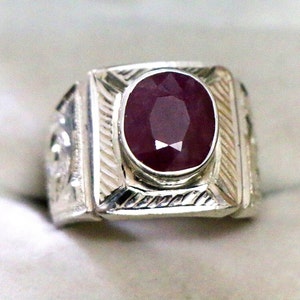 Può includere: Un anello d'argento con una pietra preziosa ovale rossa incastonata in una lunetta quadrata. L'anello ha un design dettagliato sulla fascia.