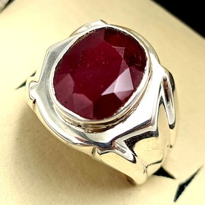 May include: A silver ring with a large, faceted, red ruby gemstone set in the center. The ring has a thick band with a textured design.