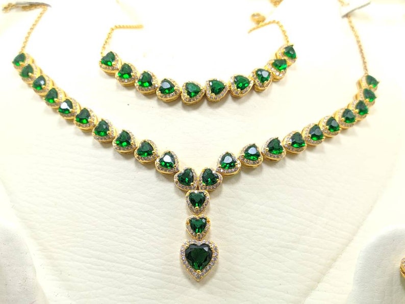 Op de afbeelding: Een gouden ketting- en armbandset met groene hartvormige stenen. De ketting heeft een hanger met een grotere hartvormige steen.