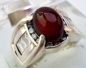 May include: A silver ring with a large, oval-shaped, dark red gemstone surrounded by smaller, clear gemstones.