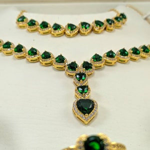 Op de afbeelding: Een gouden kettingset met een lange en een korte ketting, beide versierd met smaragdgroene hartvormige stenen omringd door kleinere heldere stenen.