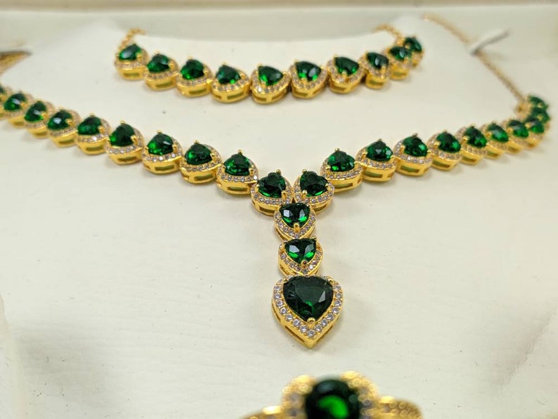 Op de afbeelding: Een gouden kettingset met een lange en een korte ketting, beide versierd met smaragdgroene hartvormige stenen omringd door kleinere heldere stenen.