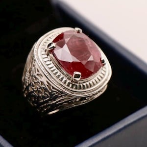 May include: A silver ring with an oval, faceted, deep red gemstone. The ring has an ornate, engraved design around the band and setting. The gemstone is the focal point, catching the light and displaying a rich color.