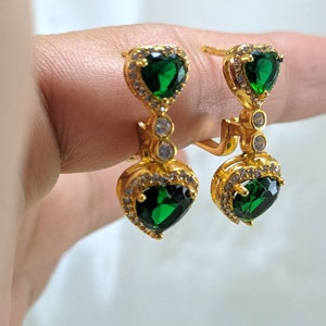 Op de afbeelding: Gouden oorbellen met een hangende groene hartvormige steen en kleinere heldere stenen.