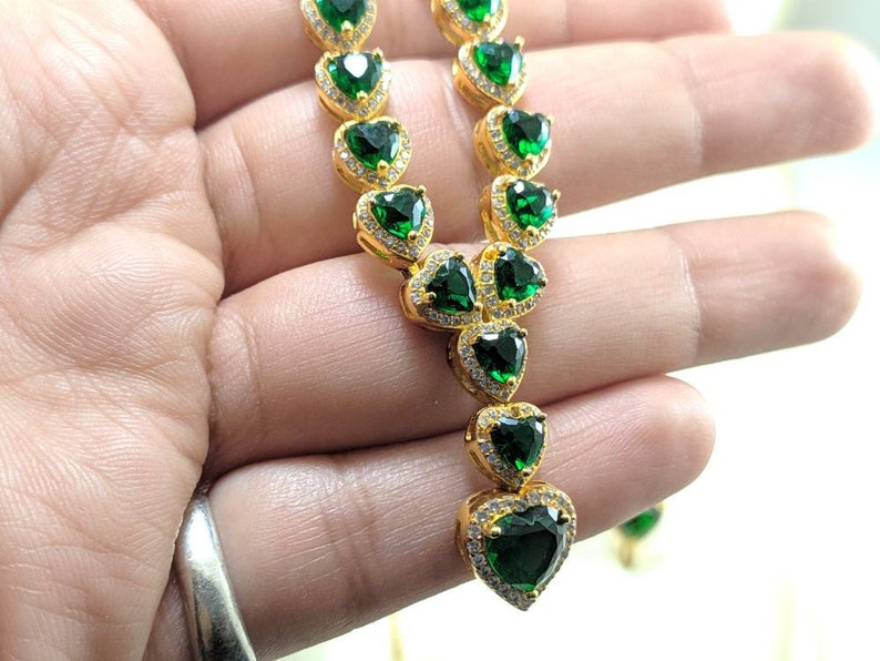 Op de afbeelding: Een gouden ketting met een hartvormige hanger. De hanger is bezet met een grote groene smaragd en kleinere heldere diamanten. De ketting is van goud en heeft een kettingschakelontwerp.