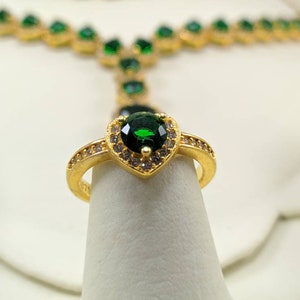 Op de afbeelding: Een gouden ring met een grote groene hartvormige edelsteen omringd door kleinere heldere edelstenen.