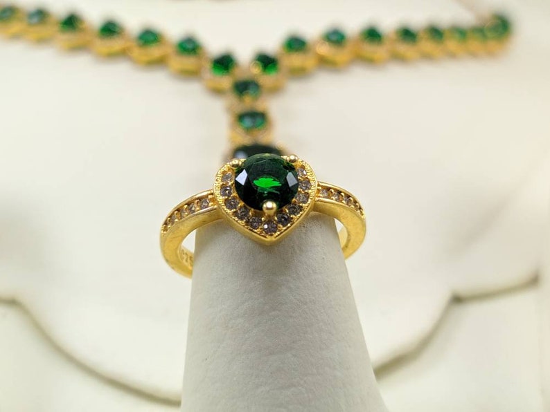 Op de afbeelding: Een gouden ring met een grote groene hartvormige edelsteen omringd door kleinere heldere edelstenen.