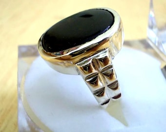May include: A silver ring with a large, oval, black stone. The ring has a polished silver band and a decorative, textured design on the sides. The stone is set in a gold-colored bezel. The ring is displayed on a white surface.