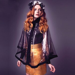 May include: A woman wearing a black and white polka dot shirt, a gold skirt, and a sheer black cape with a black trim. She is wearing a flower crown.