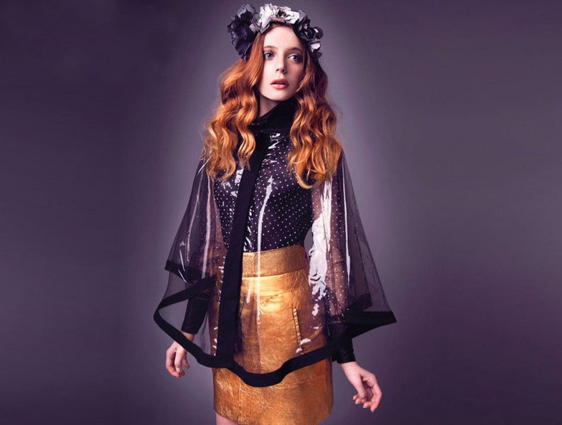 May include: A woman wearing a black and white polka dot shirt, a gold skirt, and a sheer black cape with a black trim. She is wearing a flower crown.