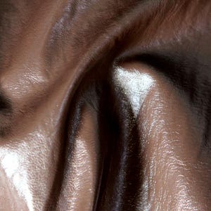 May include: A close-up view of a smooth, dark brown material with a glossy, reflective finish. The surface features deep, flowing folds and creases that create a dynamic play of light and shadow, highlighting the texture and sheen of the fabric.