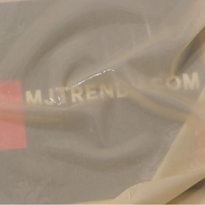May include: A translucent, off-white plastic film partially covering a dark grey square card. The card features a red rectangle on the left side and the white text MJTREND.COM printed horizontally across the centre of the dark grey background.