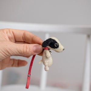 May include: A small needle-felted dog figurine with cream-colored fur, a black ear patch, and a dark eye. The dog wears a red collar with a small silver bell and is attached to a long, thin red leather strap featuring a silver metal snap button.