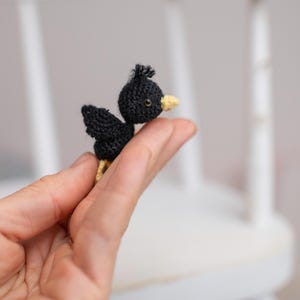 Puede incluir: Una figura de pájaro en miniatura tejida a ganchillo en color negro, con un pequeño mechón de hilo en la cabeza, un diminuto pico amarillo y un solo ojo oscuro visible. El pájaro presenta un patrón de punto texturizado y patas amarillas.