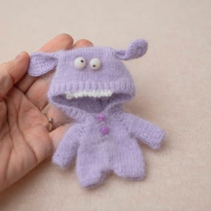 Puede incluir: Un pequeño pelele de punto color violeta claro que presenta una capucha con dos orejas caídas, dos ojos móviles blancos y una hilera de dientes blancos a lo largo de la abertura. La prenda incluye dos pequeños botones violetas en el pecho.