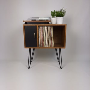 May include: A mid-century modern record player stand with a black door and open shelves for vinyl records. The stand has hairpin legs and is made of wood.