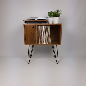 May include: A wooden record player stand with a cabinet and open shelves for storing vinyl records. The stand has black hairpin legs and is decorated with a small potted plant.
