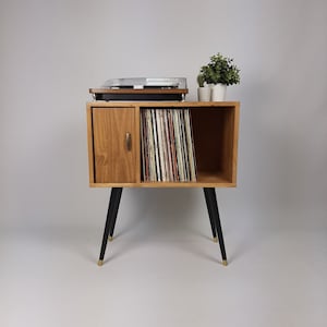 May include: A mid-century modern record player stand with a wood cabinet and black legs. The cabinet has a door on the left side and an open shelf on the right side. The shelf is filled with vinyl records. The stand has a turntable on top.