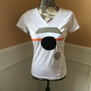 May include: White short-sleeved t-shirt with a black circle and silver and orange accents.