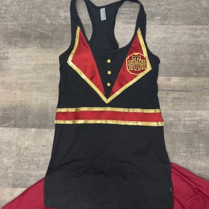 K&ouml;nnte beinhalten: Schwarzes Tanktop mit rotem und goldenem Besatz und einem goldenen gestickten Logo mit der Aufschrift "Grand Floridian". Das Tanktop wird mit einem roten Rock kombiniert.