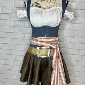 May include: A brown suede skirt with a white and red striped sash tied around the waist. The sash has a gold buckle with a silver design. The skirt is worn over a denim corset with a white blouse underneath.