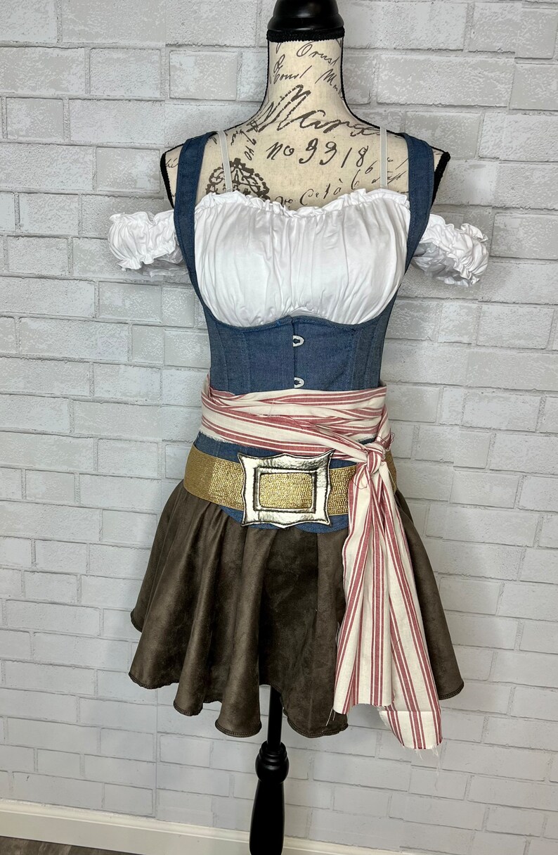 May include: A brown suede skirt with a white and red striped sash tied around the waist. The sash has a gold buckle with a silver design. The skirt is worn over a denim corset with a white blouse underneath.