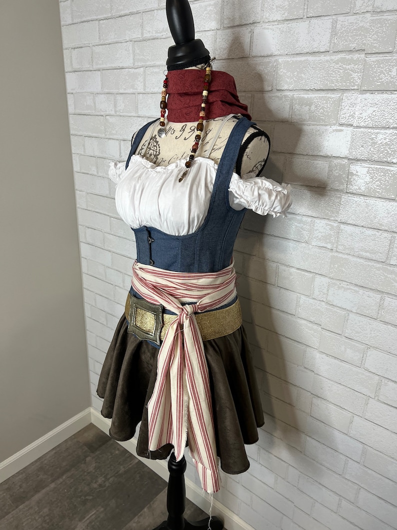 May include: A blue denim bodice with white lace trim and a brown skirt with a red and white striped sash. The bodice has a white blouse underneath and the sash is tied in a bow at the front. The mannequin is wearing a red and brown beaded necklace.